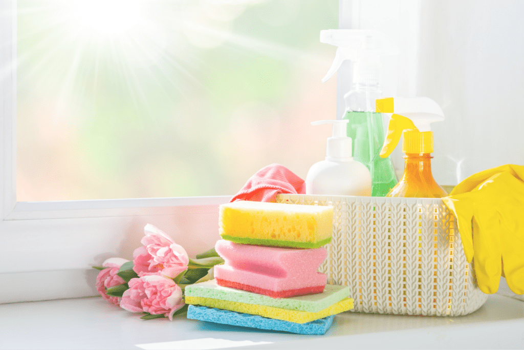 Bright colored cleaning supplies including sponges, yellow gloves, flowers, and a basket holding various spray bottles. 