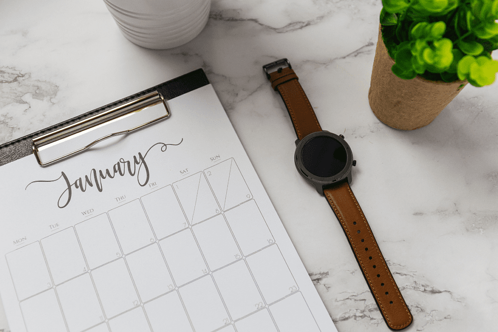 A January calendar on a clipboard on a marble counter top next to a smart watch and green plant in a brown pot. 