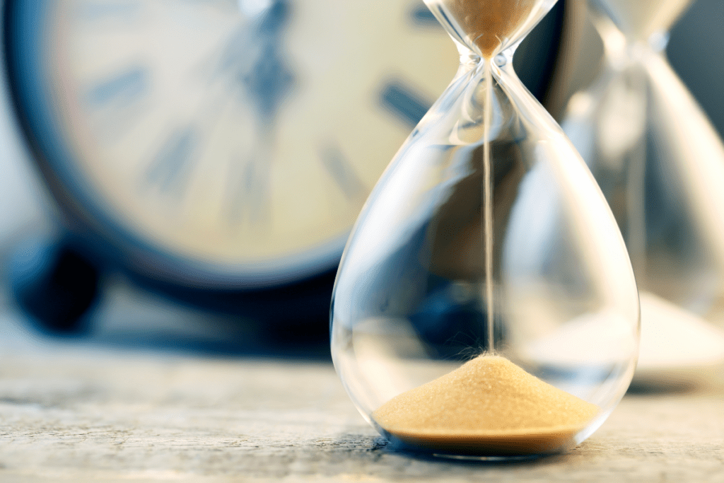 A closeup image of a the sand falling down in an hourglass with a clock blurred out in the background. 
