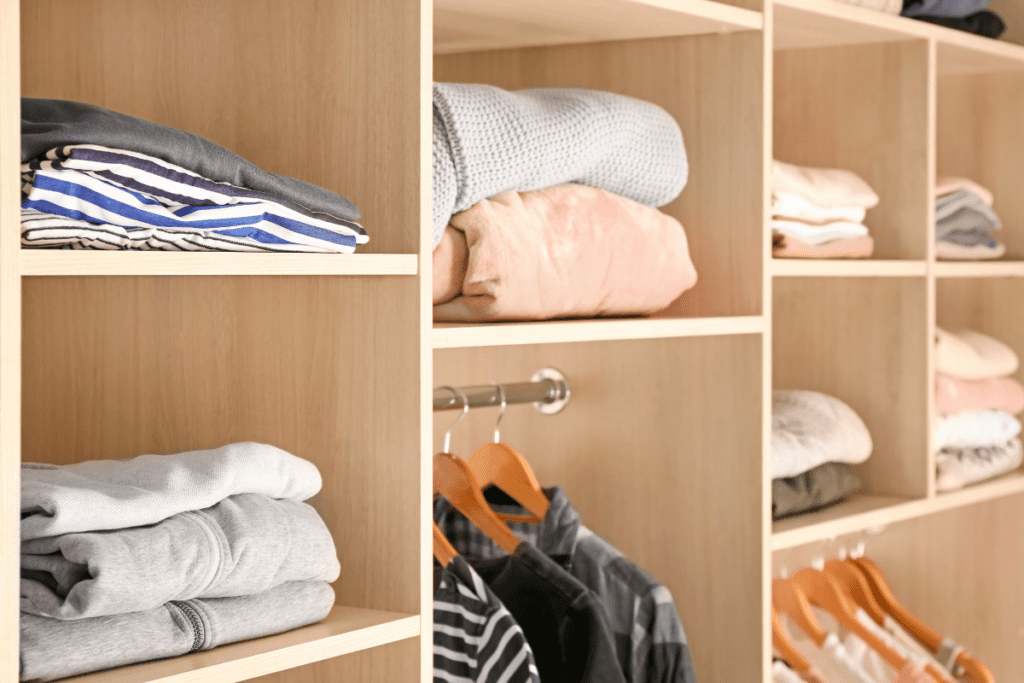 A close up image of a wooden closet organizer storing folded up clothing items stacked on top of each other and shirts hanging from wooden hangers. 