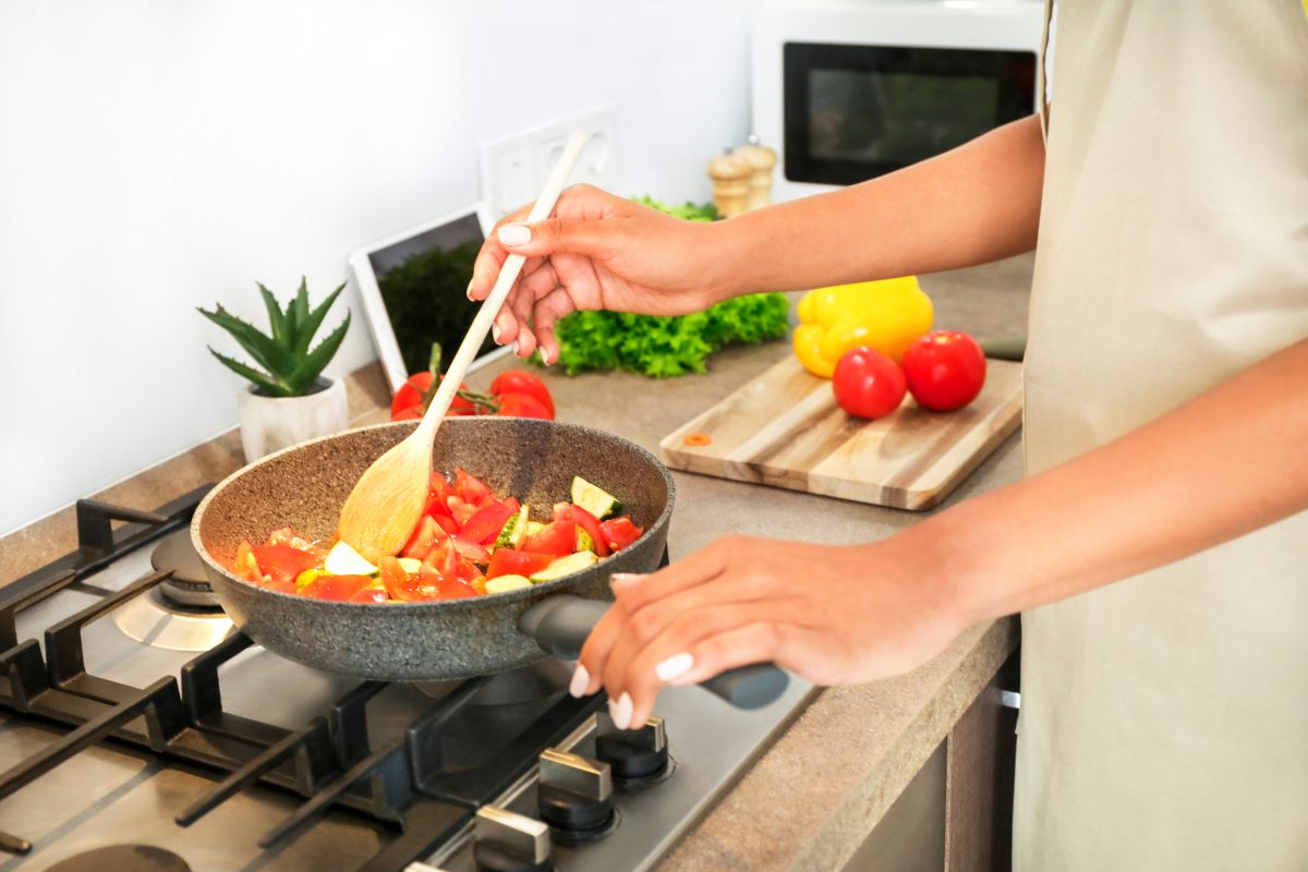 A close up picture of someone stirring vegetables in a pan on the stove with a cutting board on the counter in the background.