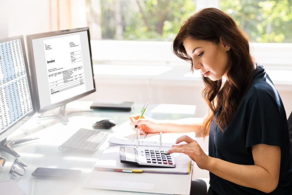 A woman sitting at a desk using a calculator with a monitor in the background showing data software.