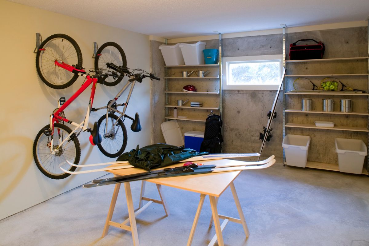 A storage area with shelves lining the walls holding plastic bins, bikes hanging on the wall, and a work table in the middle of the room with skis on it.