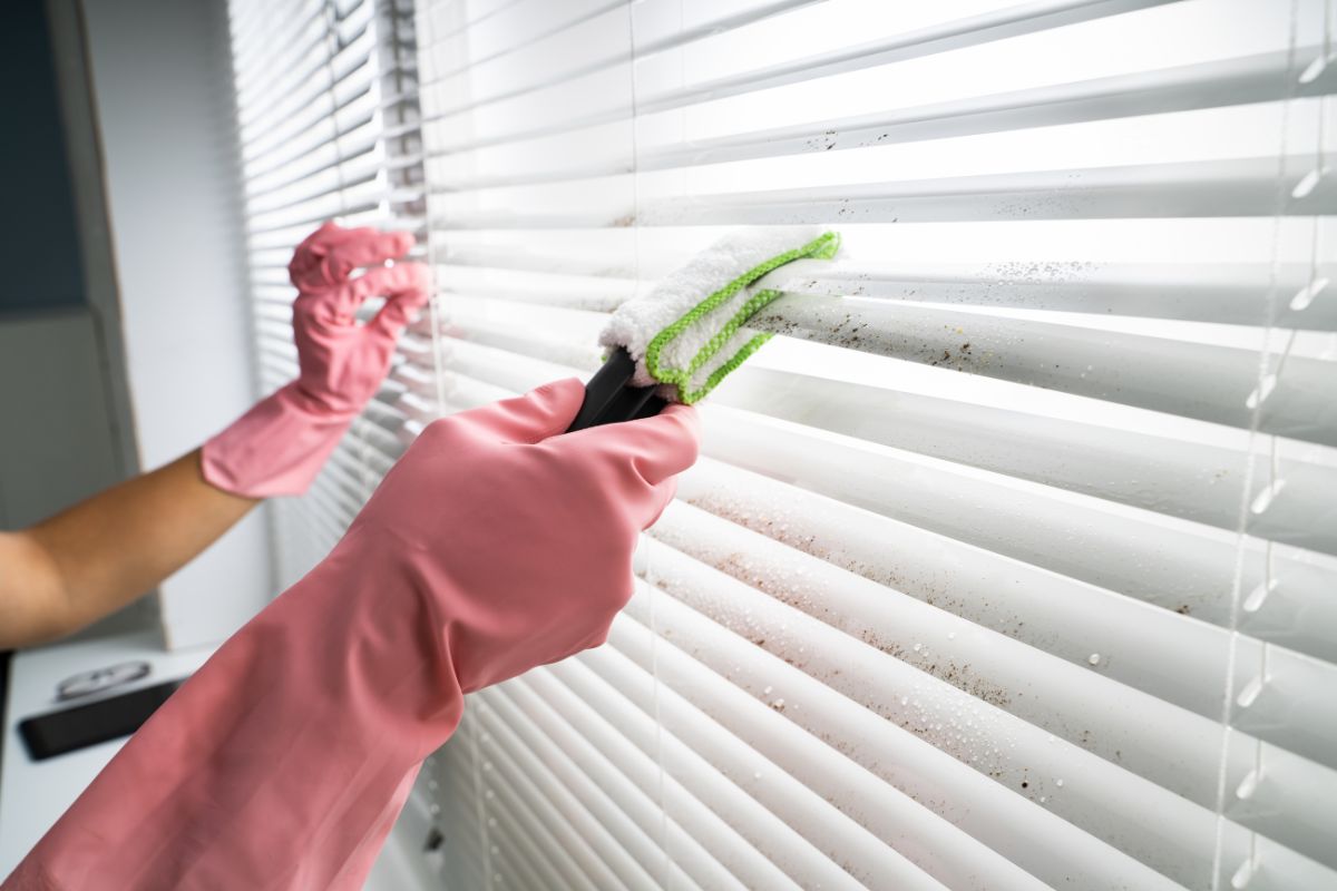 Blinds being cleaned with a microfiber tool by someone who is wearing pink cleaning gloves.