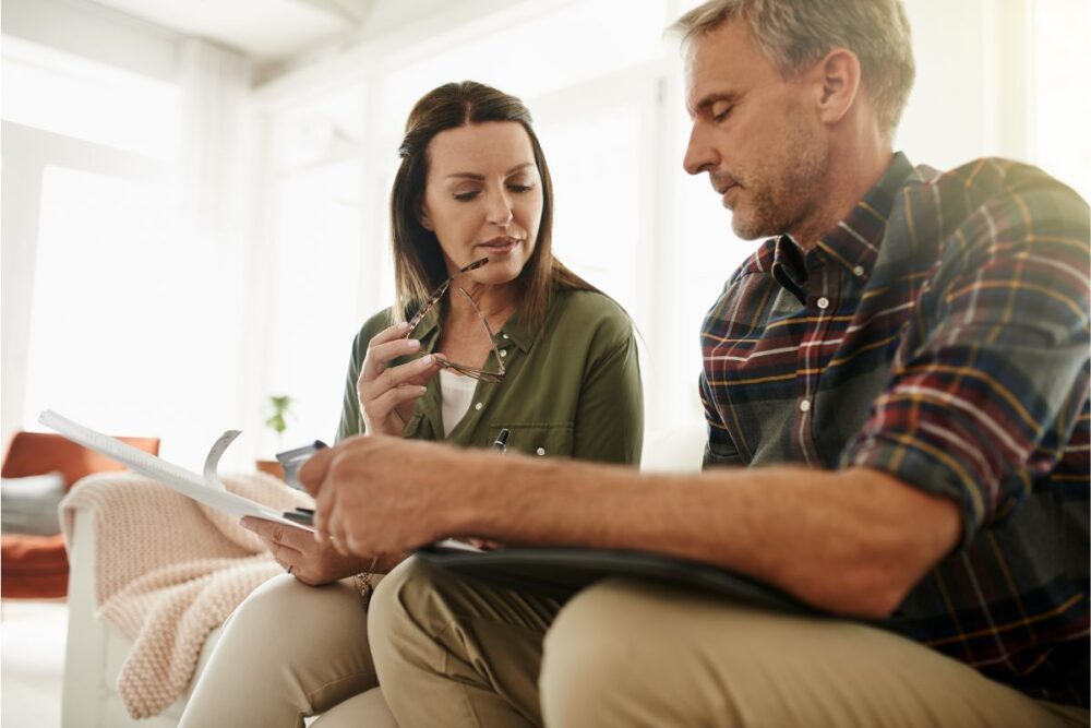 A man and woman sitting down and looking down at a pice of paper the man is holding.