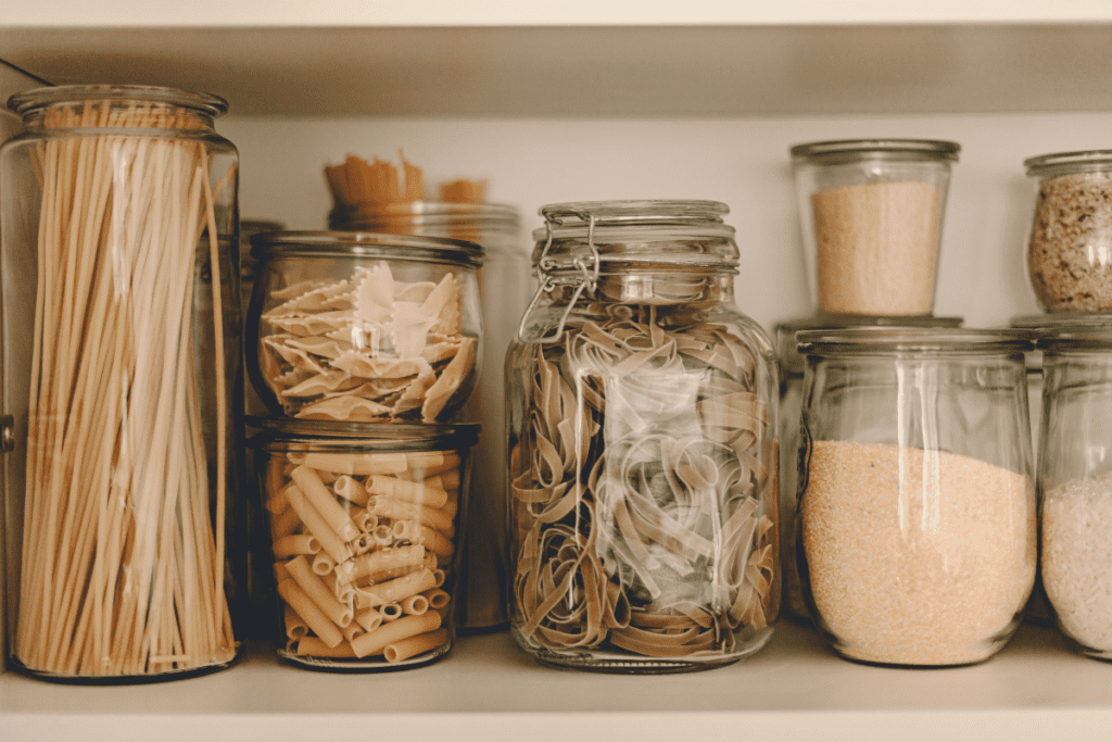 Dry food ingredients such as rice, grain, and pasta stored in clear glass containers on a shelf. 
