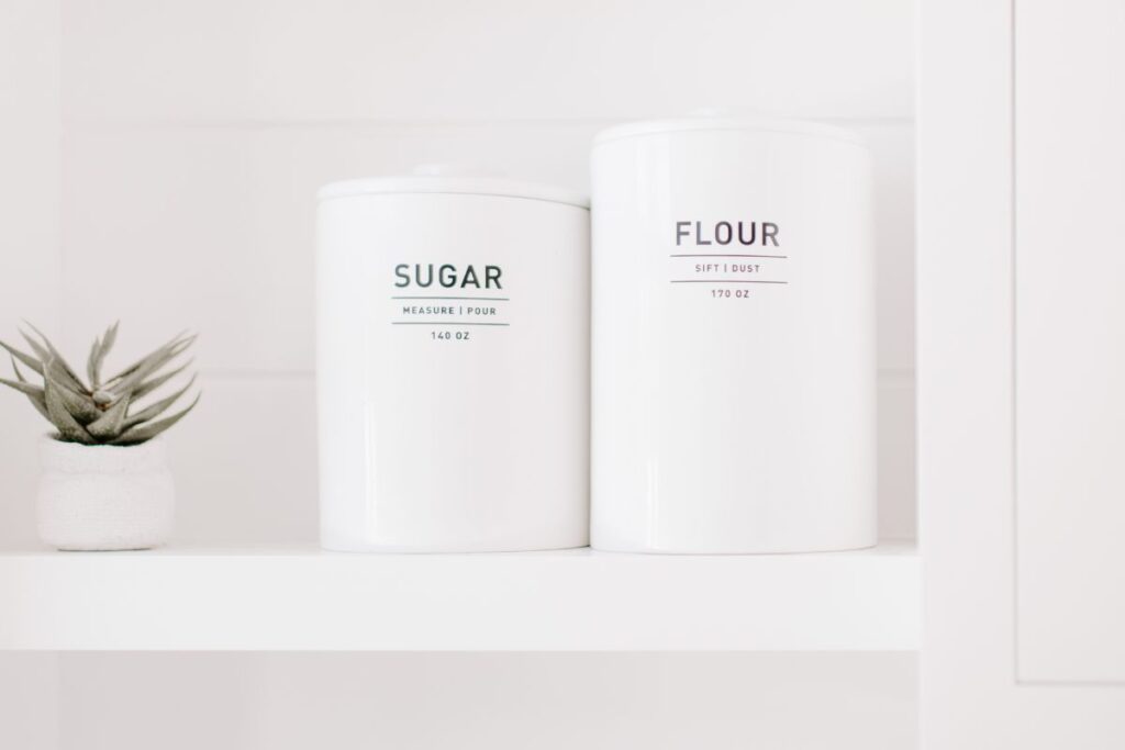 Two white canisters on a white shelf labeled "sugar" and "flour." 