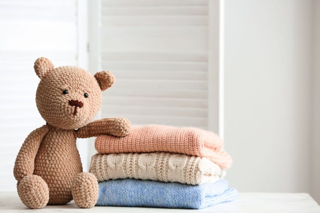 Brown teddy bear propped up with an arm leaning on a stack of three colorful sweaters.
