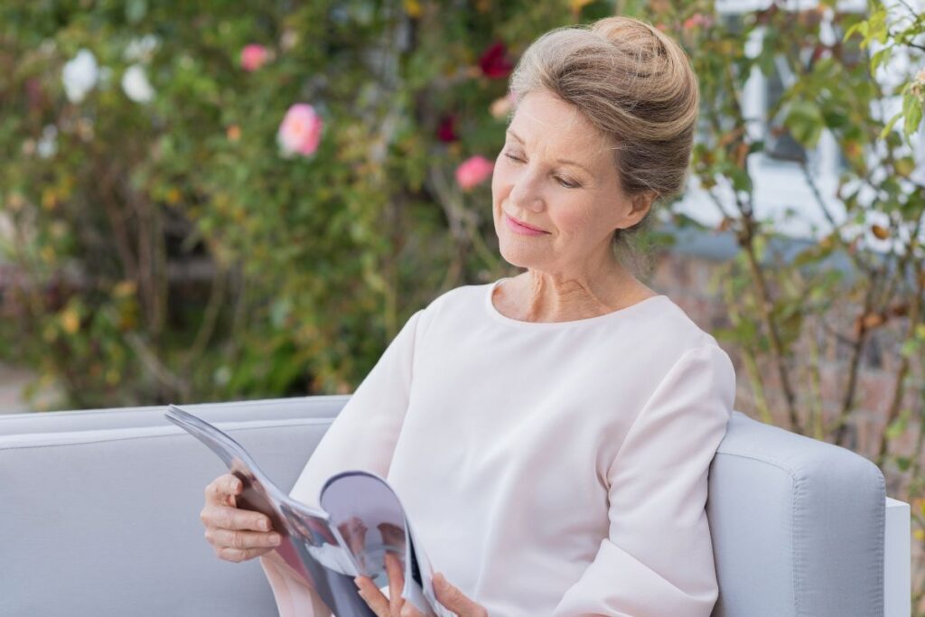 An older woman reading a magazine outside on a couch. 