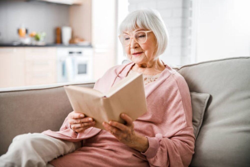 An elderly woman sitting on the couch and smiling as she reads a book.