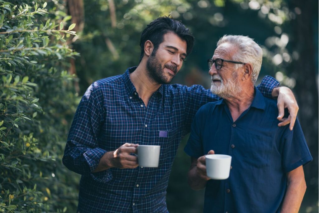 A dad and son walking with coffee mugs and the son's arm is around his dad's shoulders.