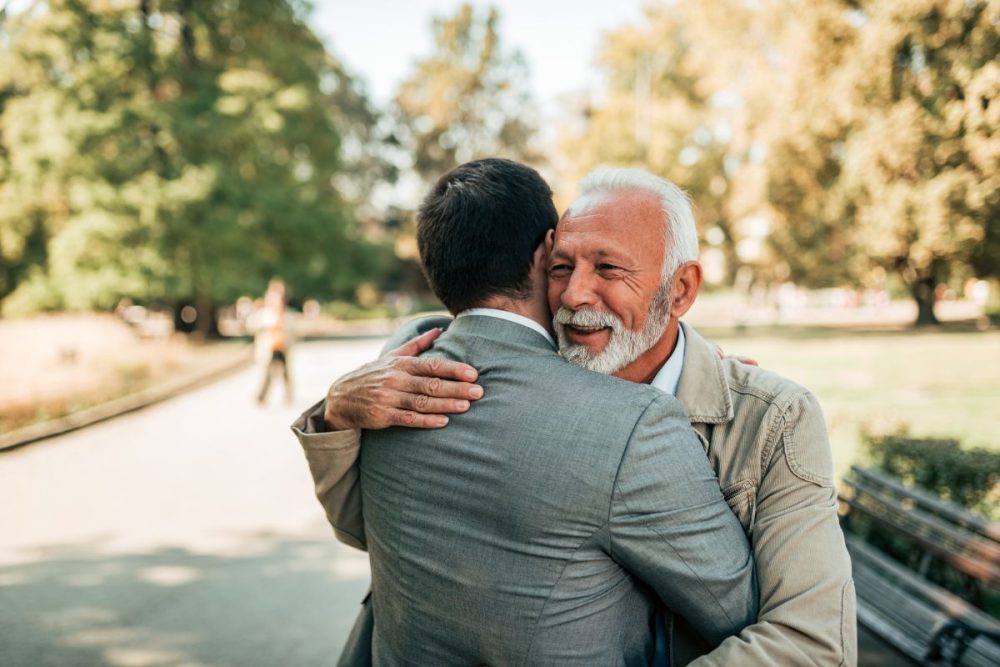 A dad giving his son a hug outside by a park bench.