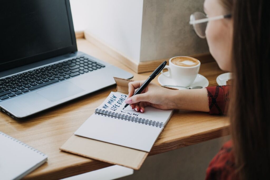 A woman sitting at a desk with a laptop, coffee, with a journal open and writing goals in an open notebook.