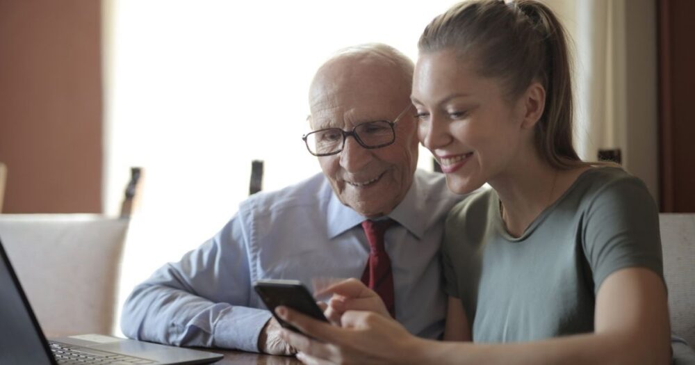 young-lady-showing-photos-on-smartphone-to-senior-man-while-sitting-at-laptop