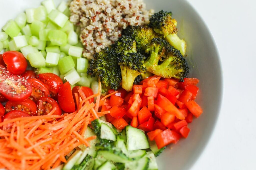 A white bowl full of healthy, colorful foods like broccoli, carrots, cucumbers, and tomatoes.