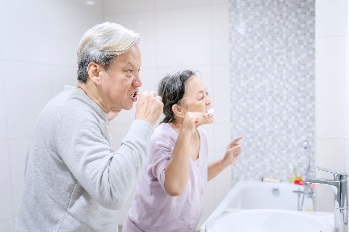 An elderly man and woman standing side by side brushing their teeth while looking in the mirror.