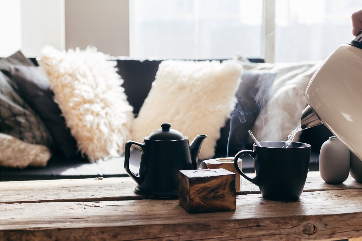 A cozy blue couch with decorative pillows behind a wooden coffee table with a black teapot and a tea cup steaming beside it.