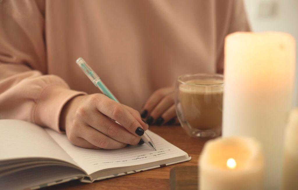 A woman in a pink sweater writing in a open journal with a blue pen with candles and a cup of coffee on the desk with her. 