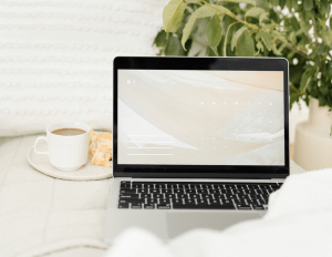 A laptop sitting on a bed with a coffee cup next to it and a plant behind it. 