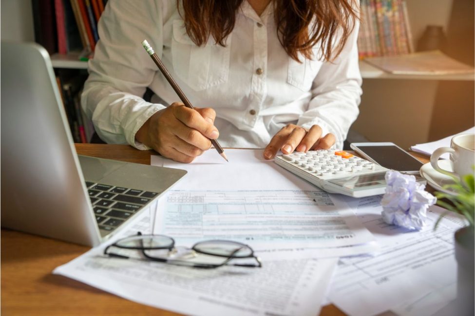 A woman sitting at a desk writing on a piece of paper with a pencil with a pair of glasses, calculator, and papers spread out.