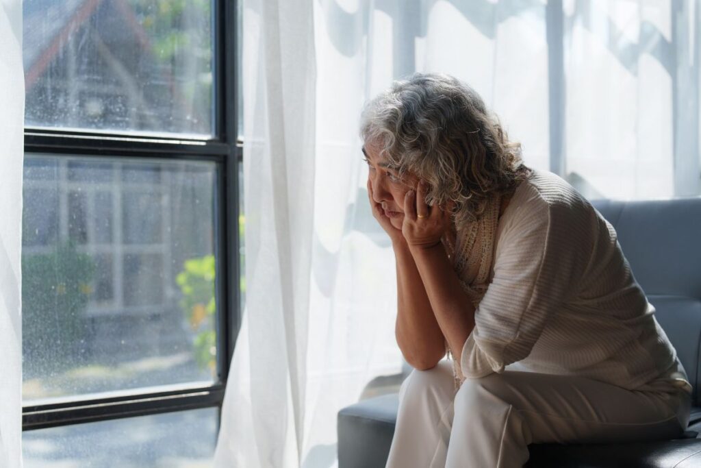 An older woman facing mental health challenges sitting on a couch with her head in her hands as she looks sadly out of a window. 