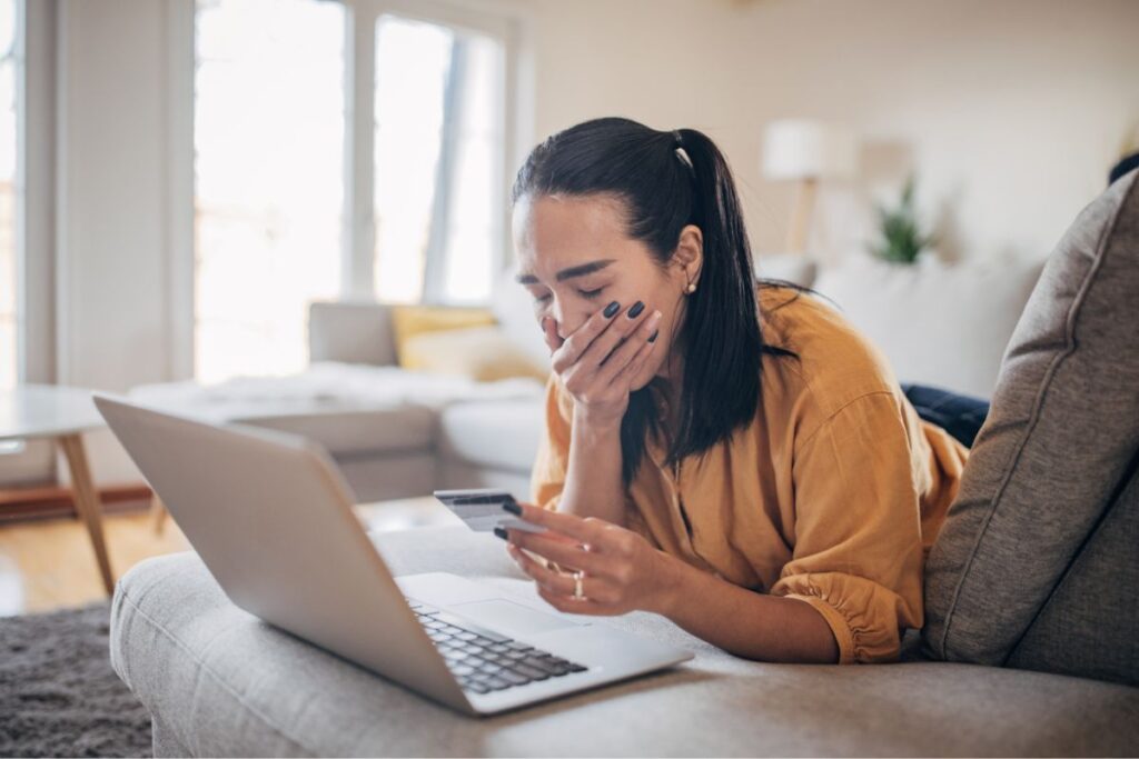 A woman laying on a couch with her laptop, holding a credit card with her hand over her mouth. 