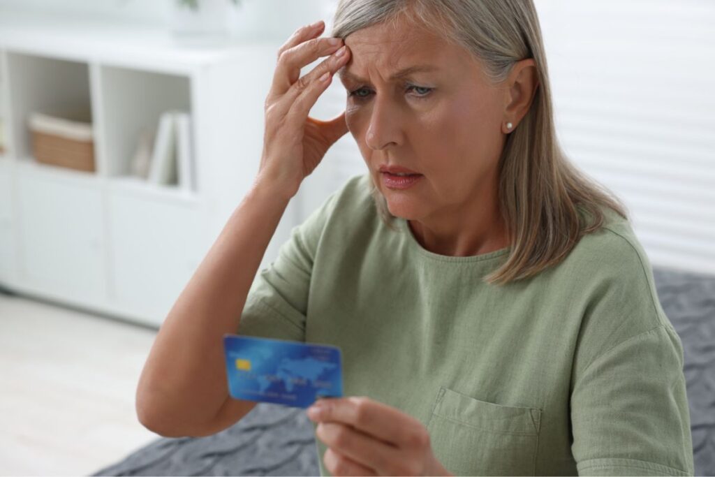 An older woman looking at her credit card with one hand resting on her head in frustration, financial scams. 