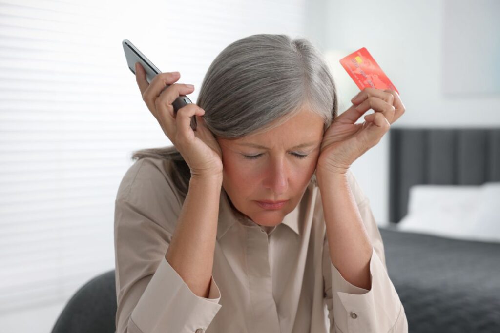 An older woman holding her head in her hands with a credit card in one hand and a phone in the other hand. 