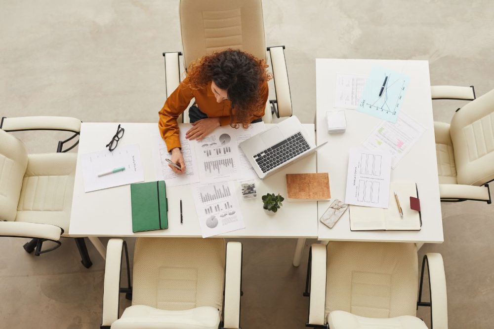 A woman sitting at a desk with papers and a laptop spread out and looking at financial documents.