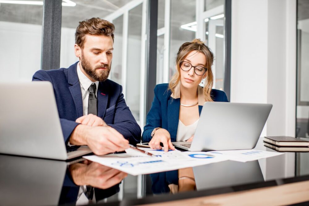 A man and a woman sitting at a desk with laptops open and looking at report documents on sheets of paper