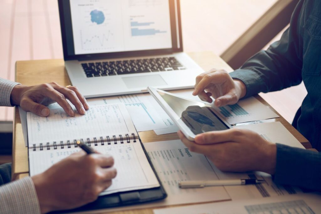 Two people sitting across from a desk with a laptop, tablet, and notebooks spread out looking a financial documents and using Simplifi by Quicken. 